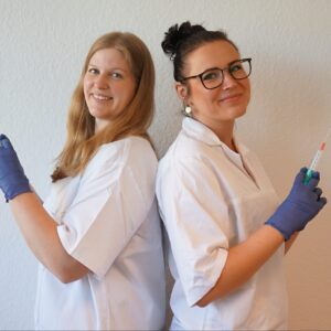 2 Personen im weißen Kasak stehen Rücken an Rücken und jeder hält eine Spritze in der Hand
